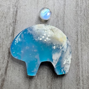 May include: A blue and white resin elephant figurine with a small, iridescent, round stone above it. The elephant has a light blue body with white cloud-like patterns. The stone has a rainbow sheen.