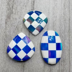 May include: Three iridescent, checkerboard-patterned cabochons in different shapes: a rounded square, a teardrop, and an oval. The squares alternate between white and blue, with a shimmering, colorful effect. The cabochons are set against a gray background.