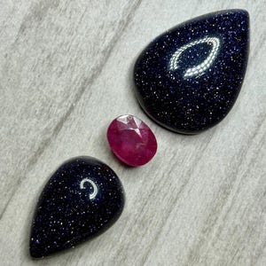 May include: Three gemstones on a light gray surface. Two large, dark blue, teardrop-shaped stones with sparkling inclusions are positioned around a smaller, oval-cut, ruby-red gemstone. The gemstones are arranged diagonally.