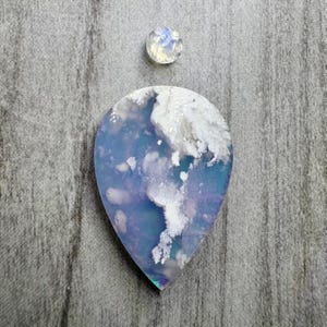 以下が含まれることがあります： 半透明の青と紫のベースに白い雲のようなインクルージョンが入ったティアドロップ型のカボションと、かすかな虹色の光沢と淡い乳白色の外観を持つ小さな丸いファセットカットの宝石のセット。