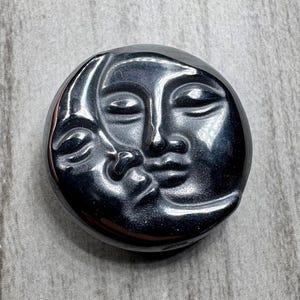 May include: A dark gray, circular decorative stone featuring a carved design of a sun and moon face. The faces are in a kissing pose, with closed eyes and detailed facial features. The stone has a polished, reflective surface.