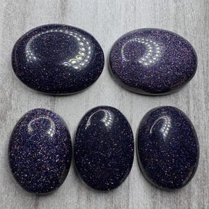 May include: Five polished blue sandstone stones with a sparkling, glittery appearance. The stones are oval and round, with a dark blue base color and flecks of gold and silver. The stones are smooth and reflective.