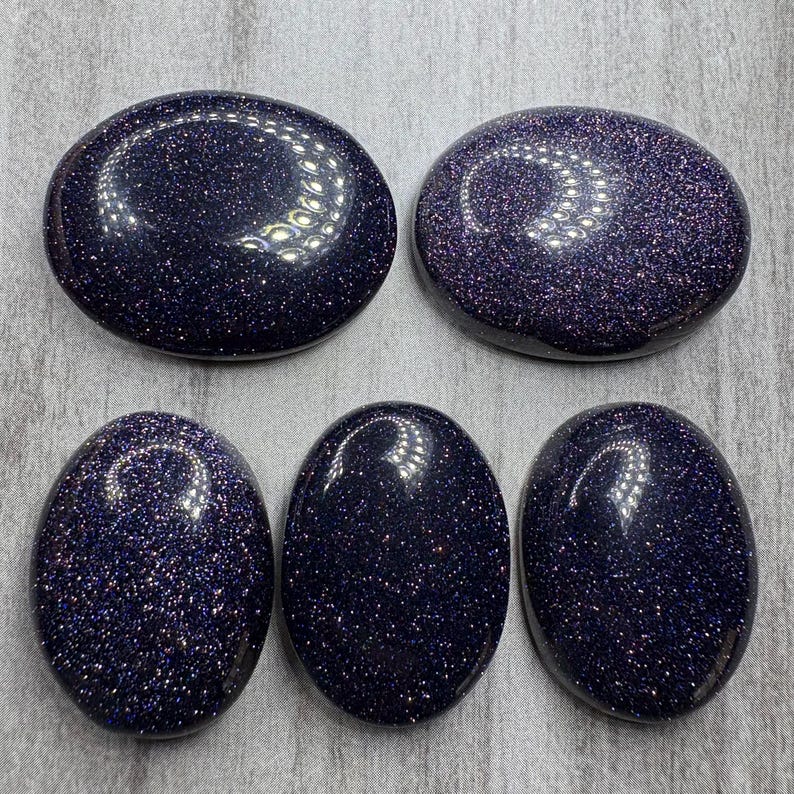 May include: Five polished blue sandstone stones with a sparkling, glittery appearance. The stones are oval and round, with a dark blue base color and flecks of gold and silver. The stones are smooth and reflective.