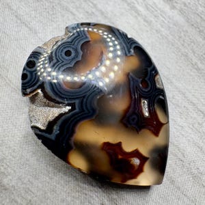 May include: A polished, teardrop-shaped agate stone featuring intricate, concentric banding in shades of dark charcoal, deep brown, and translucent amber. The surface displays a smooth, glossy finish with natural crystalline inclusions visible.