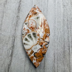 May include: A marquise-shaped gemstone with a unique pattern of white, orange, and brown hues. The polished stone has a marbled appearance with intricate details and a glossy finish. The gemstone is set against a neutral gray background.
