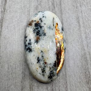 May include: Oval-shaped gemstone with a white base, speckled with black and brown inclusions. A vertical strip on the right side displays a shimmering, golden-brown iridescence. The polished stone is smooth and reflects light.