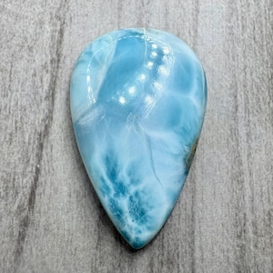 May include: A polished, teardrop-shaped gemstone with a light blue hue, reminiscent of ocean waves. The stone features swirling patterns of white and darker blue, creating a marbled effect. The surface is smooth and reflective.