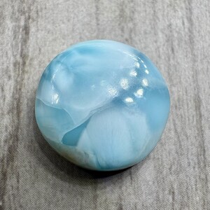 Pode incluir: Uma pedra preciosa polida e redonda, de cor azul-celeste, com padrões brancos em espiral e uma superfície lisa e refletora. A pedra apresenta tons variados de azul-claro e branco opaco, criando um aspeto marmoreado com acabamento brilhante.