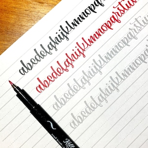 May include: A close-up of a black brush pen with a red cap, writing the alphabet in cursive on lined paper. The letters are written in black, red, and gray ink.