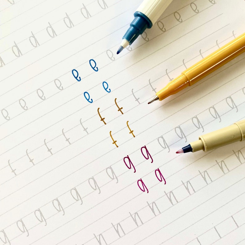 May include: A close-up of a notebook page with handwriting practice. The letters 'e', 'f', and 'g' are written in blue, orange, and purple ink, respectively. The letters are written in cursive.