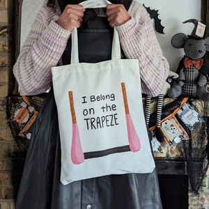May include: A white tote bag with the text "I Belong on the Trapeze" and a pink and brown illustration of a trapeze. The bag is being held by a person wearing a black dress and a pink party hat.