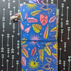 Può includere: Un quaderno blu con un motivo a foglie tropicali. Il quaderno è chiuso e ha un elastico intorno. C'è un nastro viola e un fiocco che pendono dalla parte superiore del quaderno.