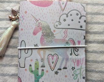 May include: A pink and white notebook with a unicorn, cactus, and stars design. The notebook has a silver elastic band and the text "You star" on the cover.