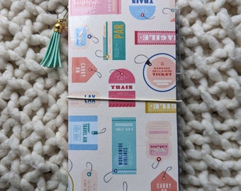 May include: A white travel journal with colourful luggage tag designs. The tags feature text like "Worldwide Airlines", "Train", "Carry On", and "Airborne".