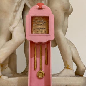 Könnte beinhalten: Eine rosa Miniatur-Standuhr mit goldfarbenem Pendel und Gewichten. Das Zifferblatt trägt den Text "Stuart Perfumes". Die Uhr ist aus Kunststoff und hat einen roten Deckel.