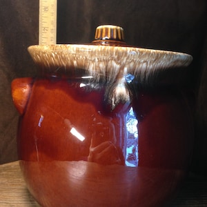 Puede incluir: Un tarro de cerámica esmaltado marrón con tapa. El tarro tiene un esmalte marrón y blanco moteado con un borde marrón. La tapa tiene un pomo marrón.