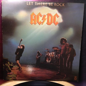 Pode incluir: Uma capa de álbum de vinil com a banda AC/DC. A capa é em preto e branco com uma foto da banda tocando no palco. O título do álbum "Let There Be Rock" está impresso em letras brancas na parte superior da capa. O nome da banda "AC/DC" está impresso em letras brancas grandes no centro da capa.