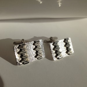 May include: A pair of silver cufflinks. Each rectangular cufflink has a textured surface with a repeating zigzag pattern in a darker silver. These cufflinks are designed for formal attire.