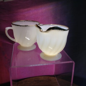 May include: A cream-coloured teacup and creamer set with gold trim. The cup has a handle and sits on a small footed base. The creamer has a spout and a similar footed base. Both pieces have a swirled design.