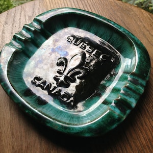 Puede incluir: Un cenicero cuadrado vintage con un esmalte verde y turquesa. El centro presenta un área plateada con las palabras "QUEBEC CANADA" y un emblema de flor de lis en negro. El cenicero tiene ranuras para apoyar cigarrillos.