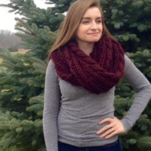 May include: A burgundy infinity scarf made of chunky knit yarn. The scarf is worn by a person standing in front of a pine tree.
