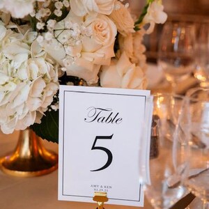 May include: A white card with black text that reads "Table 5 Amy & Ken 03.29.25" is displayed on a gold stand. The card is surrounded by white flowers.