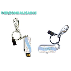 Può includere: Due portachiavi argentati con un design floreale blu e bianco. Un portachiavi ha il testo "Let's do this" e l'altro portachiavi ha il testo "Cheers".