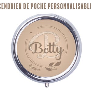 Peut inclure: Un cendrier de poche rond et argenté avec un intérieur beige. Le cendrier porte le nom "Betty" dans une écriture décorative, ainsi qu'un motif de feuilles et le texte "ATOMANIA". Le texte "CENDRIER DE POCHE PERSONNALISABLE" est en haut.