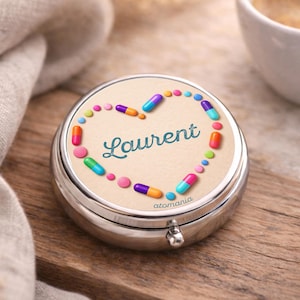 Op de afbeelding: Een zilveren pillendoosje met een beige deksel met een hartvormig ontwerp van kleurrijke pillen. De naam "Laurent" staat in turquoise letters in het hart. Op een houten oppervlak.