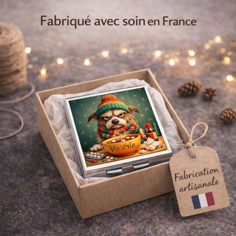 May include: A square, silver compact mirror with a colorful illustration of a grumpy dog wearing a hat and scarf, sitting in a box. The box has a tag that says "Fabrication artisanale" with a French flag.