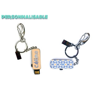 Peut inclure: Deux porte-clés argentés avec une clé USB attachée. Un porte-clés a une clé USB à motif bleu et blanc et l'autre a une clé USB beige avec le mot "Celene" imprimé dessus. Les deux porte-clés ont un anneau argenté et un clip argenté.