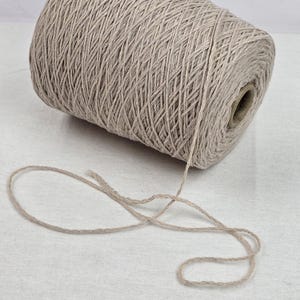 Peut inclure: Une grosse bobine de fil beige, bien enroulée, avec un brin lâche qui s'étend sur le côté. Le fil est de couleur neutre, adapté à divers projets de bricolage. Le fond est une surface blanche unie.