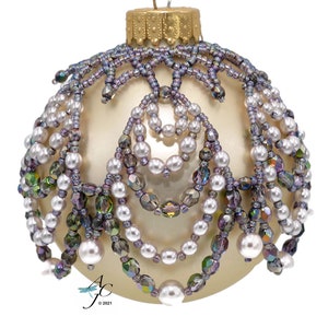 May include: Cream-colored Christmas ornament with beaded embellishments. The ornament features layers of white faux pearls and iridescent beads in shades of purple, green, and gray. A gold-colored metal cap and hanging loop complete the design.