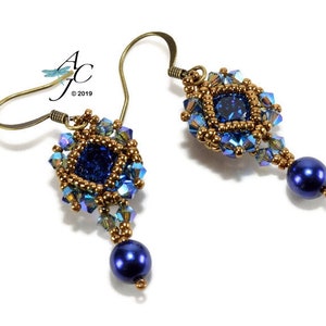 Puede incluir: Un par de pendientes colgantes con un diseño geométrico azul y dorado. Los pendientes presentan un centro de forma cuadrada con un cristal azul y están rodeados de cristales más pequeños de color dorado y azul. Los pendientes están terminados con una perla azul.