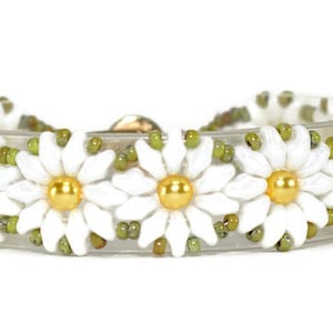 Könnte beinhalten: Ein Armband mit weißen Gänseblümchenblüten mit goldenen Zentren und grünen Perlen. Das Armband ist aus transparentem Gummiband.