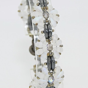May include: A beaded bracelet featuring white, petal-shaped beads and dark gray, oblong beads. The bracelet is accented with small, gold-colored beads. The design is displayed on a clear, rectangular stand. The image includes a copyright symbol and the year 2025.