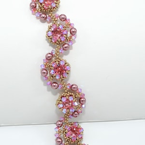 May include: A pink beaded bracelet with gold accents. The bracelet features clusters of pink crystals and pearls, connected by gold-colored beads. The clasp is gold-toned with an ornate design. The bracelet is on a white background.
