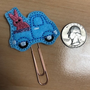 May include: A blue felt tow truck with a pink felt bunny on the back. The tow truck is attached to a gold paperclip.