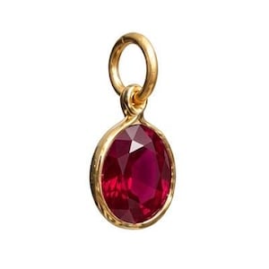 May include: A small oval-shaped pendant featuring a faceted deep red gemstone set in a polished gold-toned metal bezel. The pendant includes a circular gold-toned jump ring at the top, designed for attachment to a chain or other jewelry component.