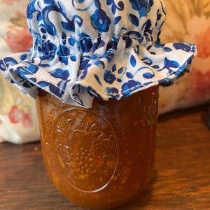 May include: A glass jar filled with a reddish-orange preserve, topped with a white fabric lid patterned with blue floral designs. The jar sits on a wooden surface, with a soft floral background.