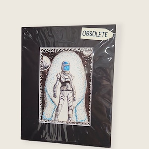 Può includere: Stampa artistica con cornice nera raffigurante un astronauta in tuta spaziale con casco blu, su sfondo azzurro chiaro e bianco. La parola "OBSOLETE" è stampata su un adesivo bianco nell'angolo in alto a destra.