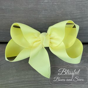 May include: A large, pale yellow hair bow made of grosgrain ribbon. The bow is centered and has multiple loops and tails. The text "Blissful Bows and Sews" is in a decorative font on the lower right.