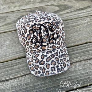 May include: A baseball cap with a leopard print pattern in shades of brown, tan, and cream. The cap features a black monogram on the front. The cap is sitting on a weathered wooden surface.