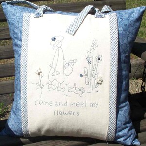 Op de afbeelding: Een decoratief kussen met een blauw-wit bloemenpatroon en een centraal paneel met een geborduurd ontwerp. Het paneel toont een grillige figuur met bloemen en de tekst "Come and meet my flowers". Het kussen heeft blauwe handvatten.