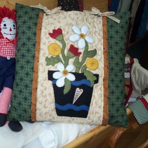 May include: A handmade pillow with a floral design. The pillow is made of fabric and features a flower pot with flowers in shades of red, white, and yellow. The pillow has a green, brown, and white patterned border. The pillow has a tag that says "Be Happy".