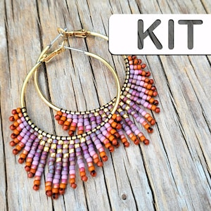 May include: Gold-coloured hoop earrings with a beaded fringe design. The fringe features a gradient of orange, purple, and pink beads. The word "KIT" is in a white rectangle in the upper right corner. The earrings are displayed on a wooden surface.