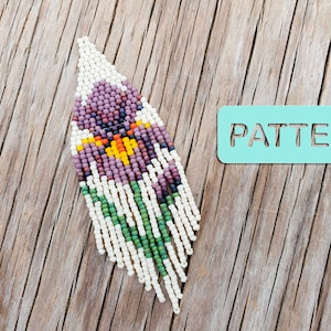 Może przedstawiać: Kolczyk z koralików z wzorem fioletowego, żółtego i zielonego kwiatu. Kolczyk wisi na drewnianej powierzchni z niebieskim napisem "PATTERN".