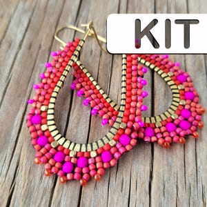 Könnte beinhalten: Ein Paar rosa und rote Perlenohrringe mit goldenen Akzenten. Die Ohrringe haben die Form einer Träne und hängen an einer Holzoberfläche. Das Wort "KIT" steht auf weißem Hintergrund in der oberen rechten Ecke des Bildes.