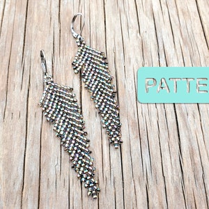 Puede incluir: Un par de pendientes largos de cuentas plateadas en forma de pluma. Los pendientes están colgados en una superficie de madera con la palabra "PATTERN" escrita en un rectángulo azul claro.