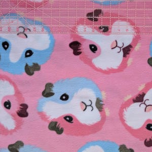 May include: Pink fabric with a repeating pattern of cartoon guinea pig faces in pink, white, blue, and black. A clear ruler with white markings is laid across the top of the fabric.